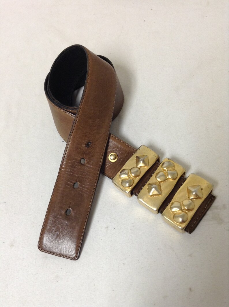 May include: A brown leather belt with a gold-toned buckle featuring multiple pyramid-shaped studs.