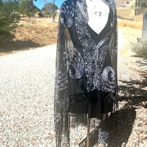 May include: A black sheer shawl with silver embroidery featuring a peacock design. The shawl has fringe along the bottom edge.