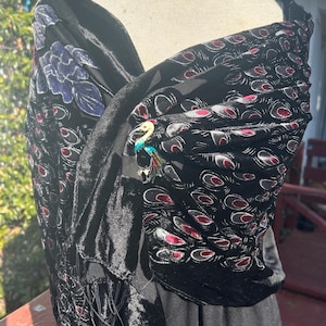 May include: Black velvet shawl with a peacock feather pattern in red and gray. A decorative brooch with a bird design is pinned to the shawl. The shawl has fringe along the bottom edge.