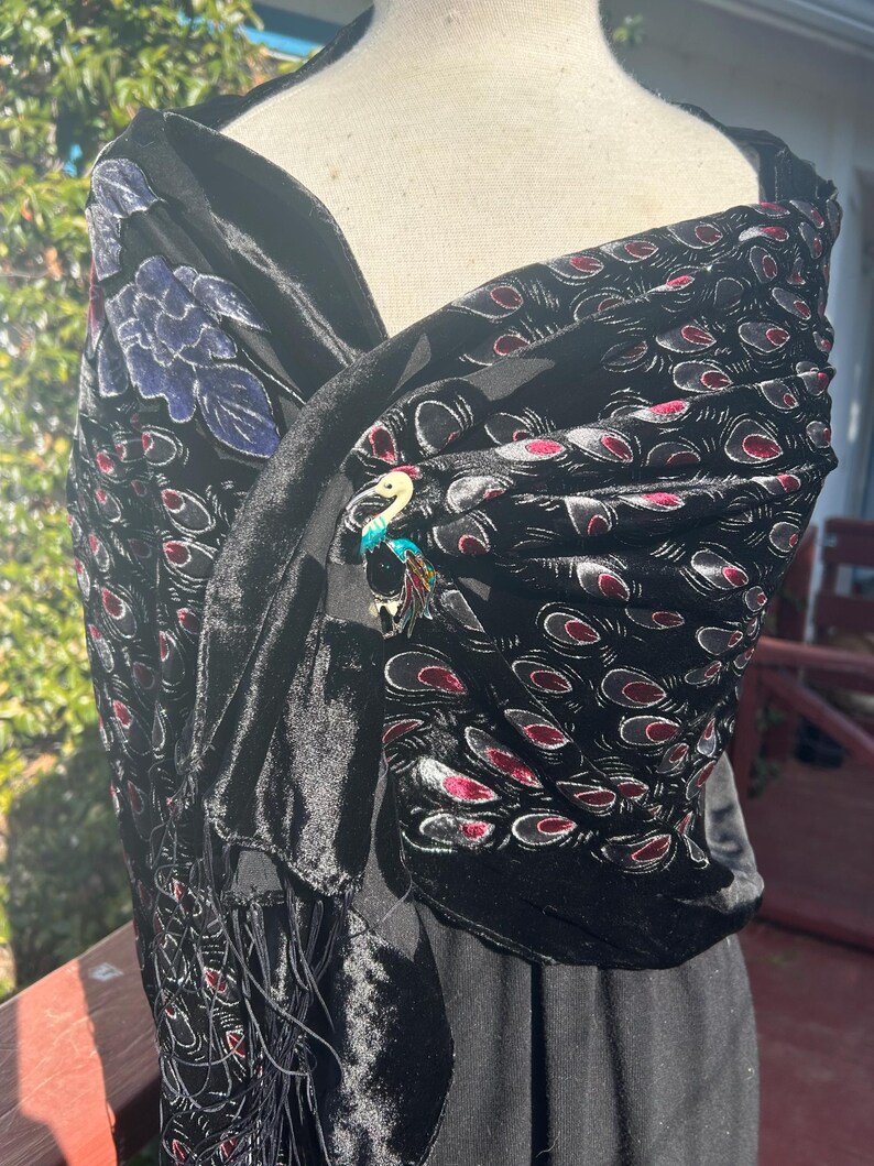 May include: Black velvet shawl with a peacock feather pattern in red and gray. A decorative brooch with a bird design is pinned to the shawl. The shawl has fringe along the bottom edge.