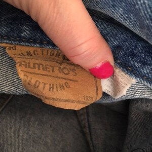 Pode incluir: Uma etiqueta de couro marrom com o texto "Junction of" "Bilmetos" "Clothing" e "1550" impresso nela.