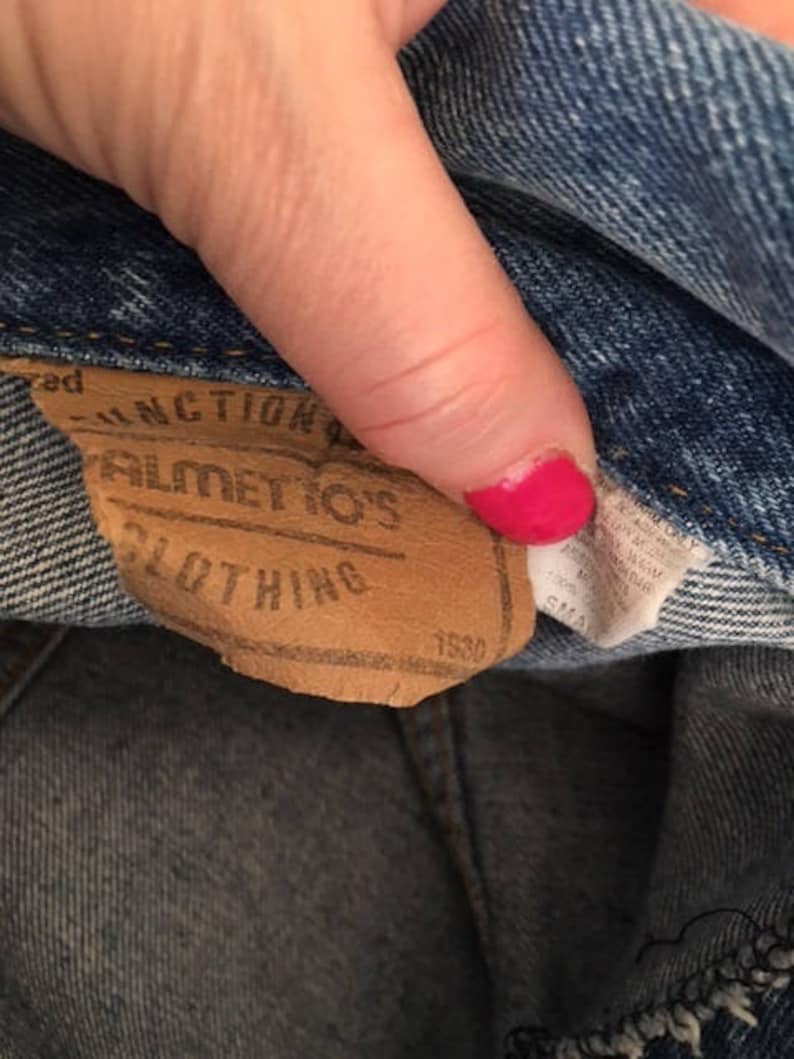 Pode incluir: Uma etiqueta de couro marrom com o texto "Junction of" "Bilmetos" "Clothing" e "1550" impresso nela.
