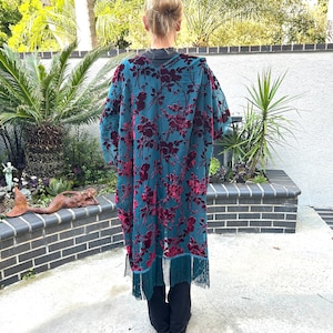 May include: A teal kimono-style robe with a floral pattern in deep red. The robe has long sleeves and a fringe trim at the hem. The person is wearing black trousers. The background includes greenery and a brick wall.