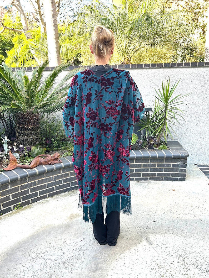 May include: A teal kimono-style robe with a floral pattern in deep red. The robe has long sleeves and a fringe trim at the hem. The person is wearing black trousers. The background includes greenery and a brick wall.