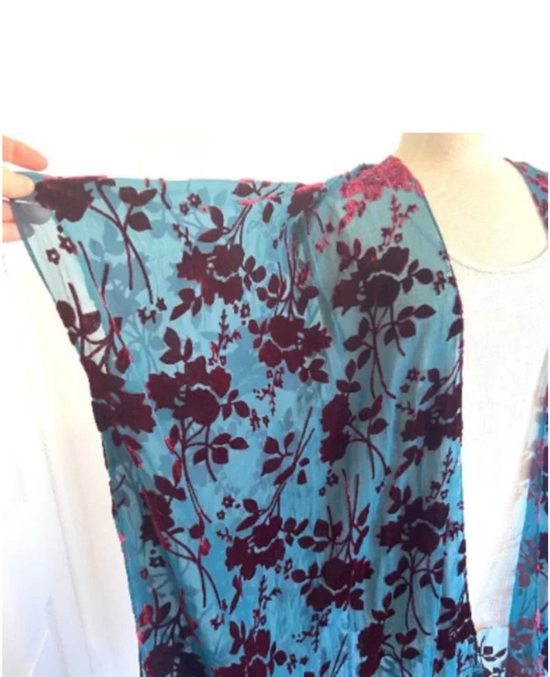 May include: A light blue kimono with a floral pattern in deep burgundy. The flowers and leaves are densely arranged across the fabric. The edges of the garment are trimmed with a darker, textured material.