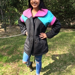 May include: Black, color-blocked jacket with a pink collar and turquoise and pink accents. The jacket has a drawstring waist and is worn with blue jeans and black boots. The jacket is a retro style.
