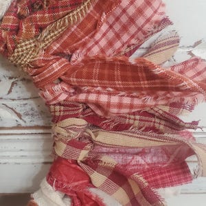 May include: A close-up of a handmade fabric wreath with frayed edges. The wreath features various red, pink, and beige plaid and patterned fabrics, tied together. The background is a white, distressed wooden surface.