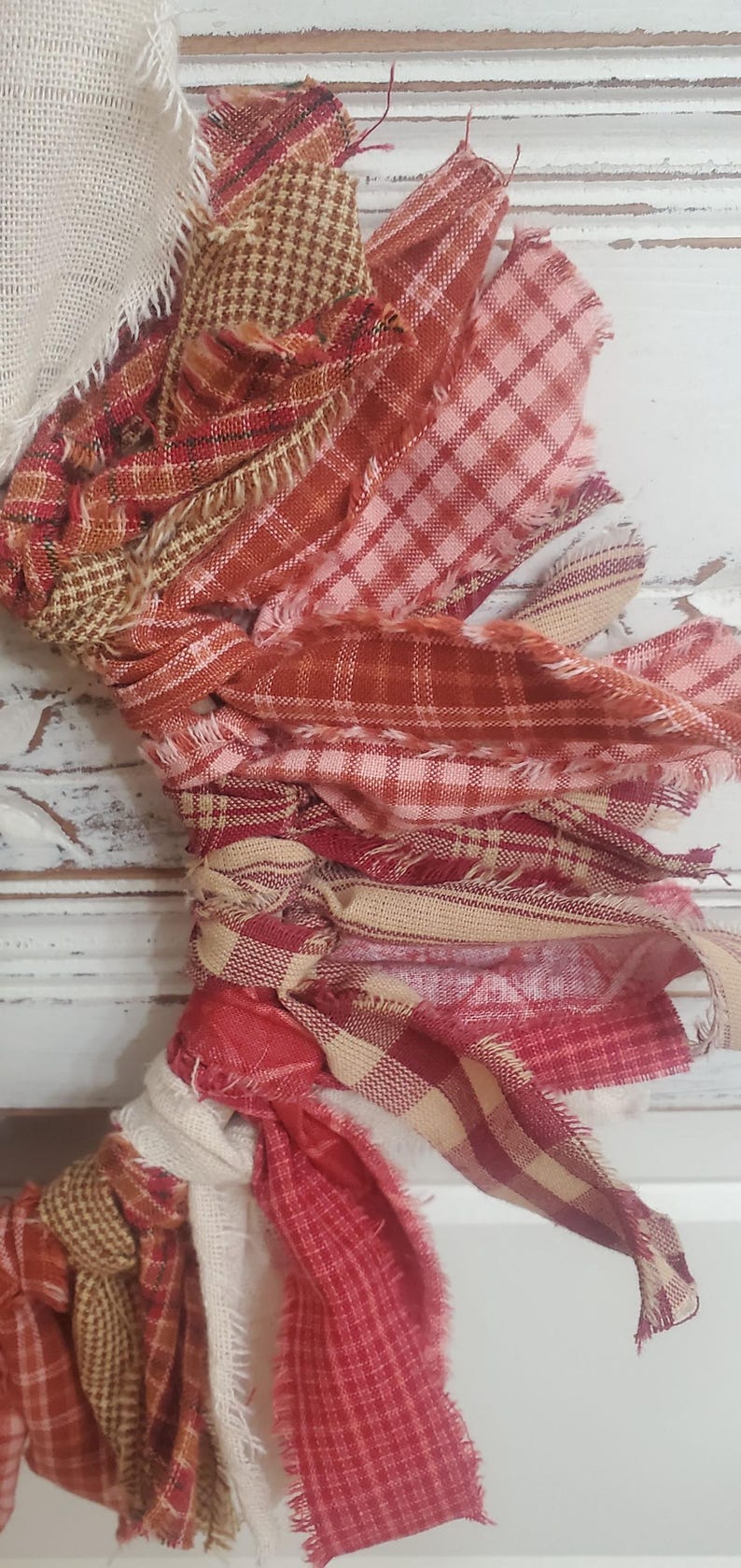 May include: A close-up of a handmade fabric wreath with frayed edges. The wreath features various red, pink, and beige plaid and patterned fabrics, tied together. The background is a white, distressed wooden surface.