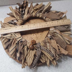 May include: A brown and tan fabric wreath with a wooden base and two small brown bells. The wreath is made of strips of fabric that have been frayed and tied together.