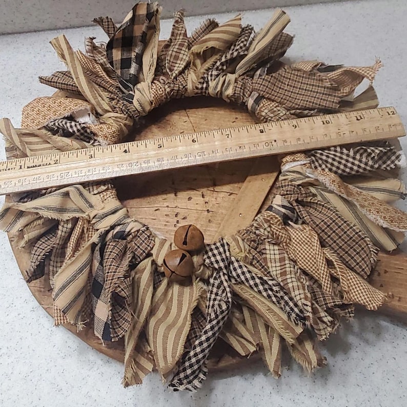 May include: A brown and tan fabric wreath with a wooden base and two small brown bells. The wreath is made of strips of fabric that have been frayed and tied together.