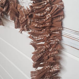 May include: A brown fabric garland with a plaid pattern, hanging from a dark metal hook. The garland is made of knotted strips of fabric, creating a rustic, textured appearance. The background is a white wall with a decorative trim.