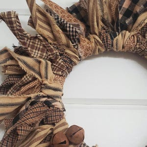 May include: A brown and tan fabric wreath with black and white plaid accents. The wreath is decorated with two small brown bells.