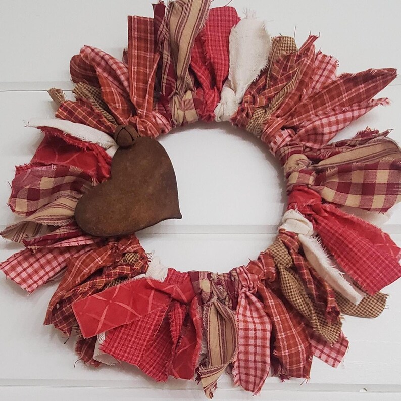 May include: A handmade wreath featuring a heart-shaped accent. The wreath is composed of various red, cream, and tan fabric strips in plaid and solid patterns. The heart is a dark brown color. The wreath is a decorative item.