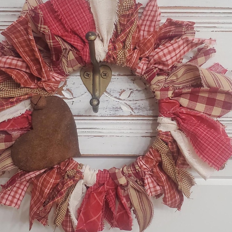May include: A handmade wreath with a heart-shaped accent. The wreath is crafted from various red and cream-colored fabric scraps, including plaid and striped patterns. A large, brown heart is attached to the wreath, which hangs on a brass hook.