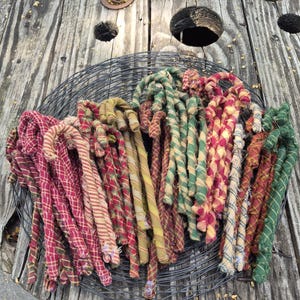May include: A collection of fabric-wrapped candy cane ornaments featuring various plaid and striped patterns in shades of red, green, yellow, and tan. The candy canes are arranged in a circular black wire basket with a thin, frayed textile texture.