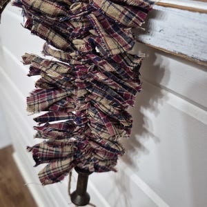 May include: A rustic, plaid fabric garland in shades of burgundy, navy, and tan. The fabric is frayed, giving a homespun appearance. The garland is attached to a dark metal rod with twine accents, suitable for seasonal decor.
