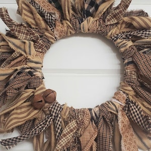 May include: A brown and tan fabric wreath with frayed edges and two small brown bells. The wreath is made of various plaid and striped fabrics.