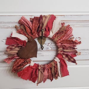 May include: A handmade wreath with red, cream, and brown fabric strips. A rusty brown heart-shaped ornament hangs from the wreath, which is adorned with a brass hook. The wreath is a rustic, country-style decoration.