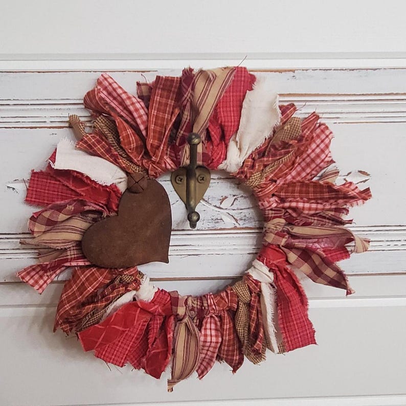 May include: A handmade wreath with red, cream, and brown fabric strips. A rusty brown heart-shaped ornament hangs from the wreath, which is adorned with a brass hook. The wreath is a rustic, country-style decoration.
