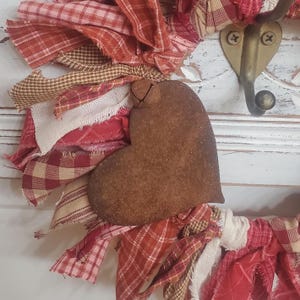 May include: A rustic, heart-shaped metal ornament with a textured, brown finish. It is adorned with a small bell and surrounded by fabric strips in red, cream, and tan plaid patterns, creating a farmhouse aesthetic.