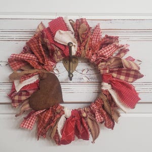 May include: A handmade fabric wreath with red, white, and tan fabric strips. A brown heart-shaped accent is attached to the wreath. The wreath is hanging on a white door with a decorative hook.