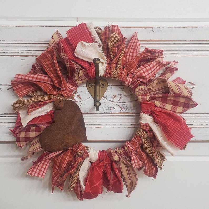 May include: A handmade fabric wreath with red, white, and tan fabric strips. A brown heart-shaped accent is attached to the wreath. The wreath is hanging on a white door with a decorative hook.