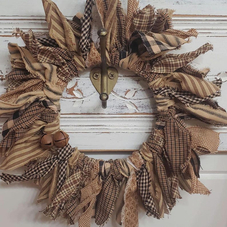 May include: A brown and tan fabric wreath with frayed edges, decorated with small bells. The wreath is hanging on a white door with a metal hook.