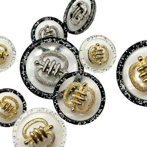 以下が含まれることがあります： 黒と銀のグリッターのアクセントが付いた、丸い白と透明なボタンの詰め合わせ。各ボタンの中央には、金または銀の金属のデザインが施されています。