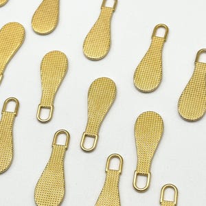 May include: Ten gold zipper pulls with a textured surface. The pulls are shaped like a teardrop with a small loop at the top.