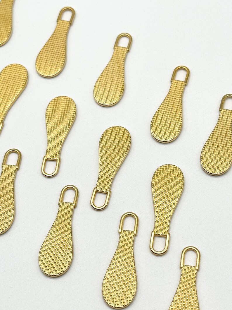 May include: Ten gold zipper pulls with a textured surface. The pulls are shaped like a teardrop with a small loop at the top.