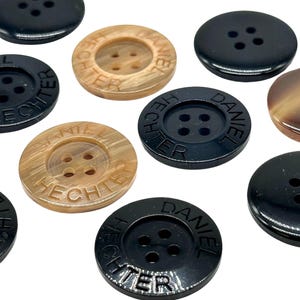 Könnte beinhalten: Verschiedene runde Knöpfe in Schwarz und Beige. Einige Knöpfe tragen die Gravur "DANIEL HECHTER". Jeder Knopf hat vier Löcher zum Annähen. Die Knöpfe bestehen aus glänzendem Material.