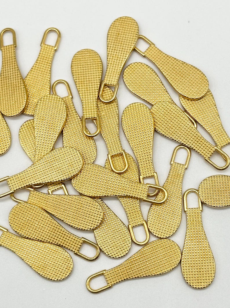 May include: A pile of gold-colored zipper pulls with a textured surface. The pulls are shaped like teardrops with a loop at the top.