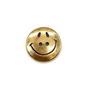 Pu&ograve; includere: Un bottone sorridente color oro con due fori per cucire. Il bottone &egrave; rotondo e ha una finitura lucida.