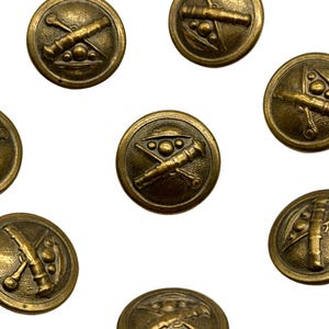 Puede incluir: Un conjunto de diez botones de metal dorado con un diseño en relieve de cañones cruzados y una corona. Los botones son redondos y tienen una superficie lisa.
