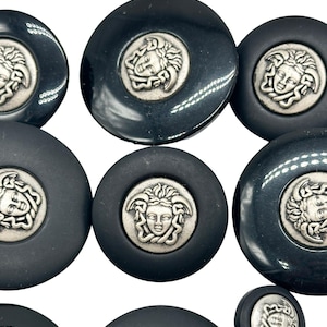 Könnte beinhalten: Ein Satz von acht schwarzen und silbernen Knöpfen mit einem Medusa-Kopf-Design. Die Knöpfe sind rund und haben einen erhabenen, silberfarbenen Medusa-Kopf in der Mitte.