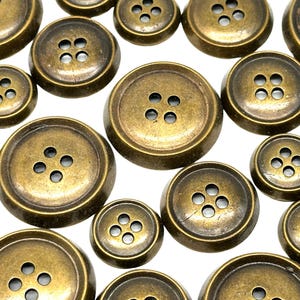 May include: Close-up of various sizes of antique brass buttons with four holes. The buttons have a slightly domed shape and a worn, vintage appearance. The buttons are scattered on a white background.