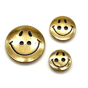 Pu&ograve; includere: Tre bottoni a forma di smiley face dorati con contorni neri. I bottoni sono di dimensioni diverse.