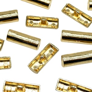 Op de afbeelding: Een verzameling goudkleurige metalen knopen in twee vormen: rechthoekig en cilindrisch. De rechthoekige knopen hebben een verhoogd midden en twee gaten aan elke kant. De cilindrische knopen zijn glad en hebben een licht afgeronde rand.