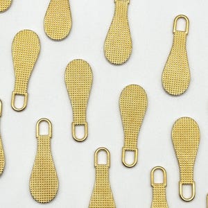 May include: A group of ten gold zipper pulls with a textured surface. The zipper pulls are in the shape of a teardrop.