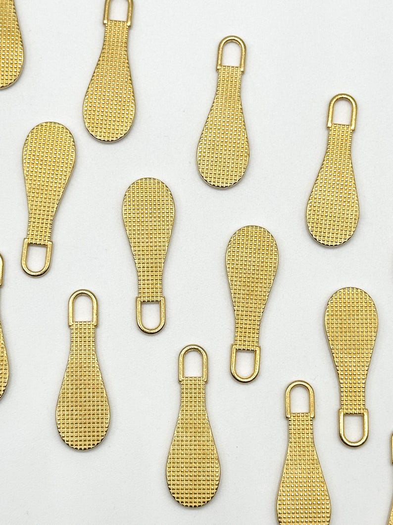 May include: A group of ten gold zipper pulls with a textured surface. The zipper pulls are in the shape of a teardrop.