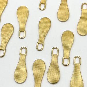 May include: A set of 12 gold zipper pulls with a textured surface. The pulls are shaped like a teardrop and have a small loop at the top.