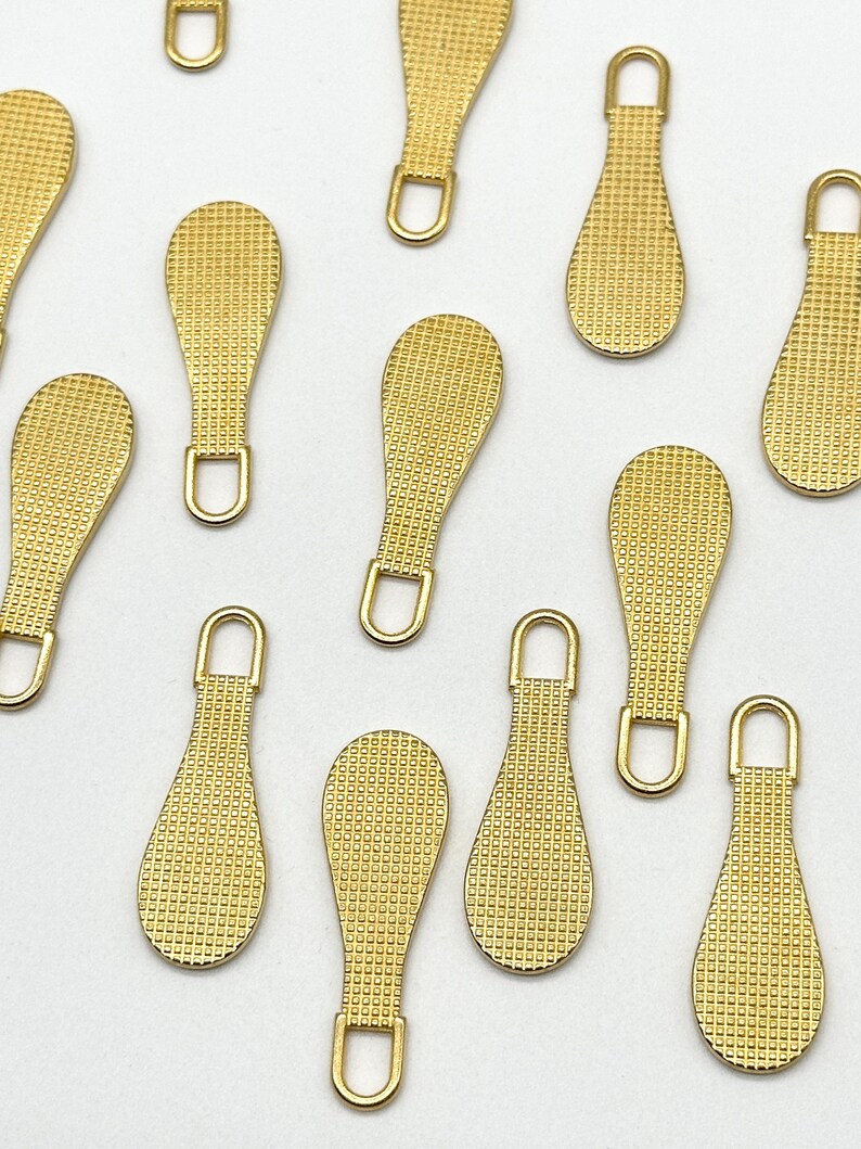May include: A set of 12 gold zipper pulls with a textured surface. The pulls are shaped like a teardrop and have a small loop at the top.