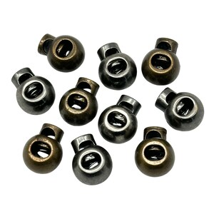May include: Ten antique gold and silver toggle stoppers for cords or drawstrings. The stoppers have a round shape with a hole in the center.