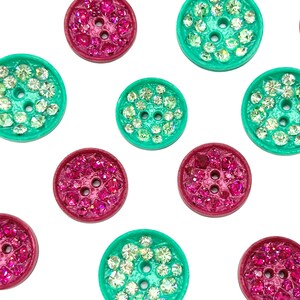 May include: Close-up of several decorative buttons. The buttons are round with a flat surface and have two or four holes for sewing. Some buttons are a bright teal colour, and others are a deep magenta. The button surfaces are covered in small, sparkling rhinestones.