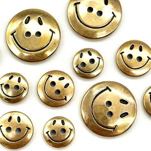 Pu&ograve; includere: Assortimento di bottoni con faccine sorridenti dorati con contorni neri. I bottoni sono di varie dimensioni.