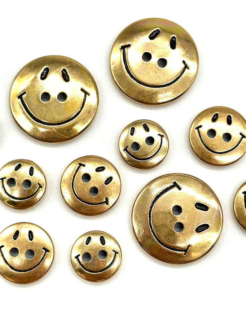 Pu&ograve; includere: Assortimento di bottoni con faccine sorridenti dorati con contorni neri. I bottoni sono di varie dimensioni.