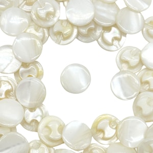 May include: A collection of white, round, mother-of-pearl buttons. The buttons have a smooth, shiny surface and a slightly iridescent sheen.