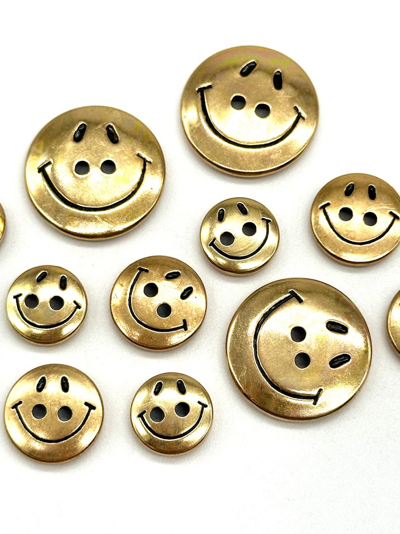 Pu&ograve; includere: Assortimento di bottoni con faccine sorridenti dorati con contorni neri. I bottoni sono di varie dimensioni.