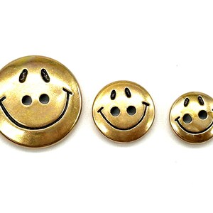 Pu&ograve; includere: Tre bottoni a forma di faccina sorridente dorati di diverse dimensioni. I bottoni hanno contorni neri e due occhi neri.