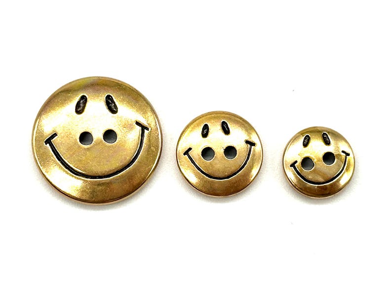 Pu&ograve; includere: Tre bottoni a forma di faccina sorridente dorati di diverse dimensioni. I bottoni hanno contorni neri e due occhi neri.